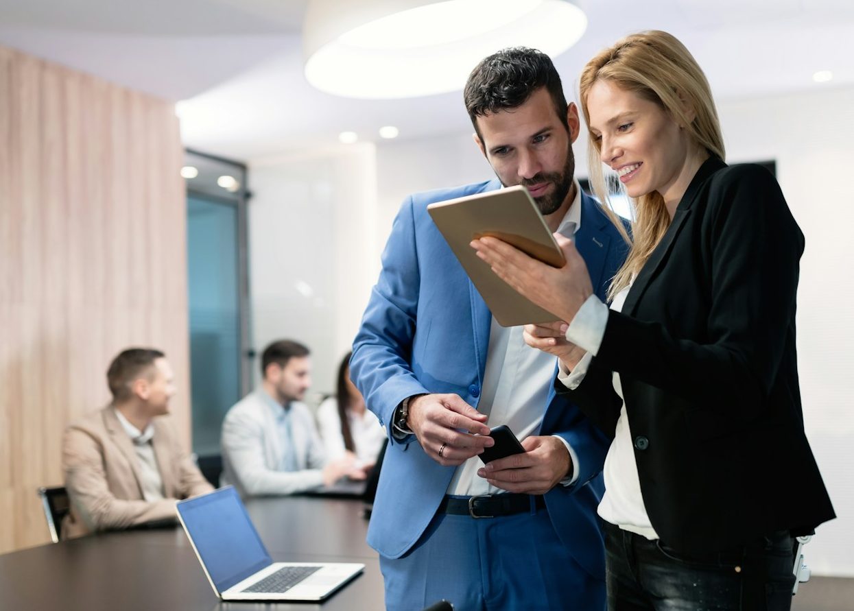 Zwei Personen befinden sich im Gespräch und blicken dabei auf ein Tablet. Im Hintergrund sitzen weitere Teammitglieder an einem Tisch.