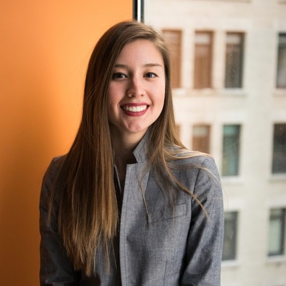 Eine lächelnde Frau mit langen Haaren im Business Outfit steht vor einer orangenen Wand. Im Hintergrund sieht man den Blick aus dem Fenster.