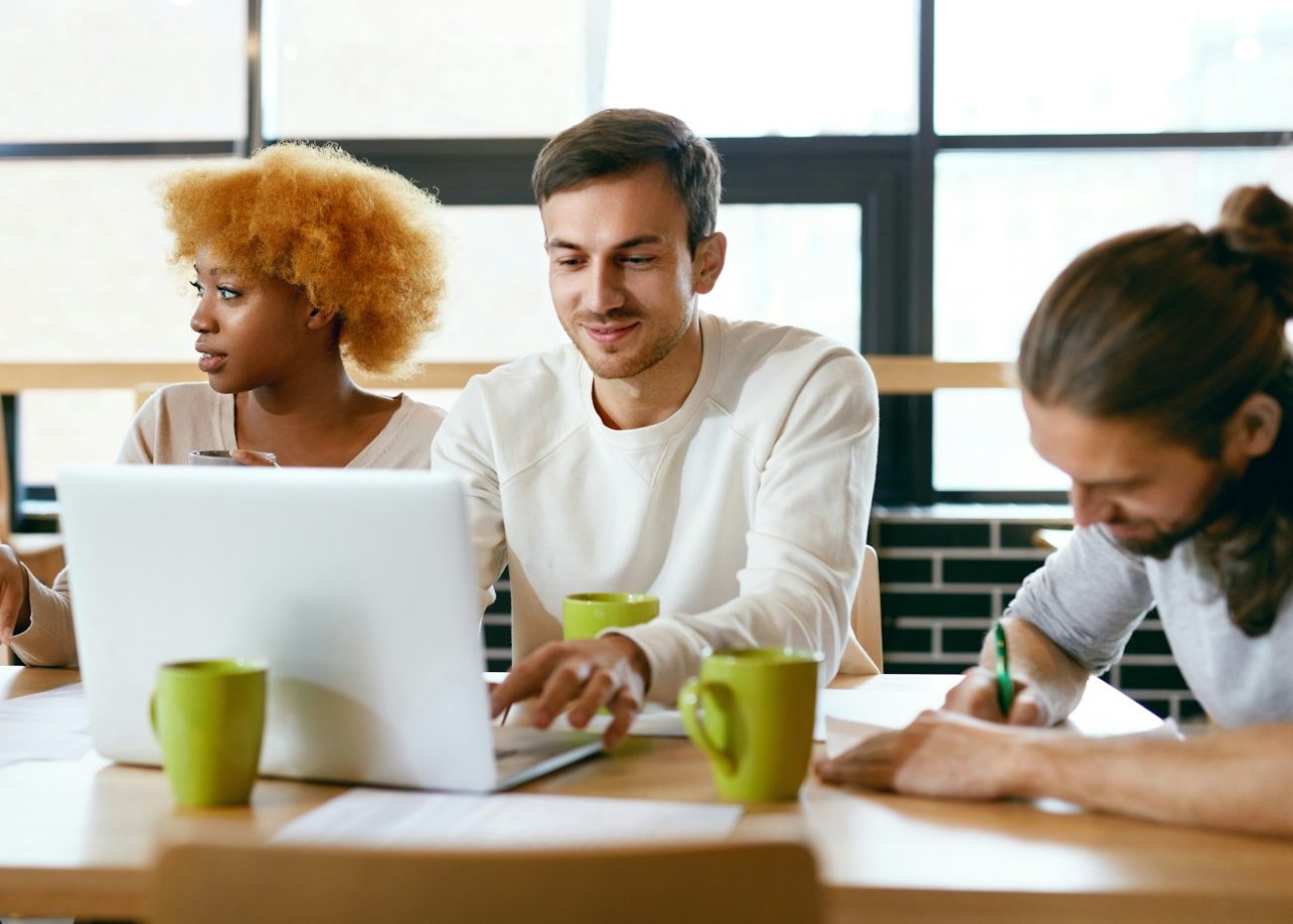 Drei glückliche Personen arbeiten gemeinsam an einem Schreibtisch. Ein Mann blickt in sein Notebook. Drei Kaffeetassen sind zu sehen.